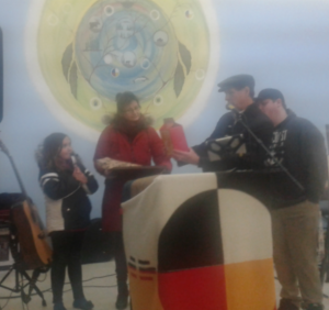 A woman and child are presented with a gift by a man with a man behind him, a podium sits in front of them covered by a quilted blanket, a colourful mural is shown on the wall in the background.