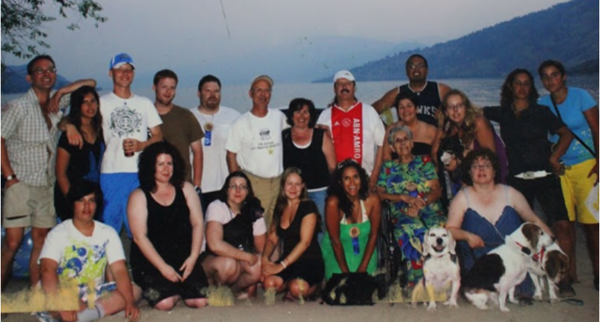 A large group of people stand together on a sandy beach for a photo with a lake shown in the background.