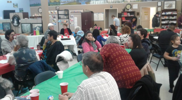 Men, women and children sit at tables in a room, some holding red cups in their hands.