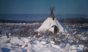 A snow-covered teepee sits in the middle of a field with a wired fence showing behind and forested hillside in the background.