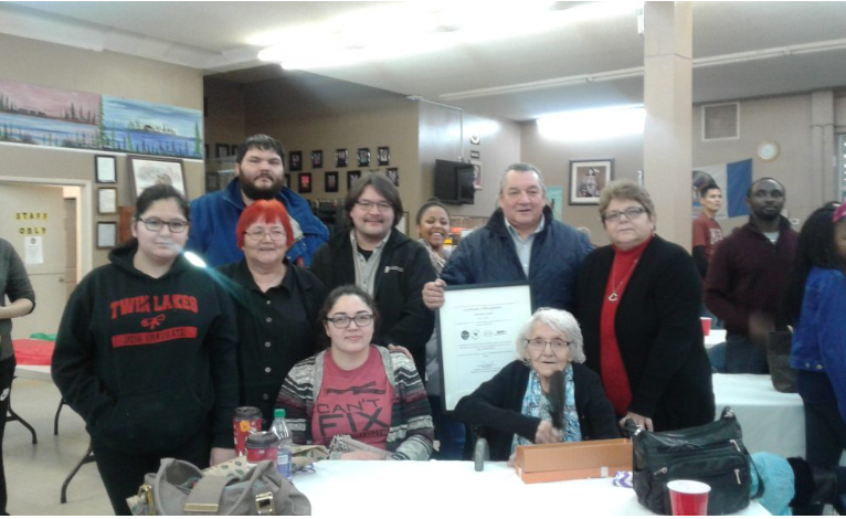 An elderly Kay Loutitt sits down at a table with her grown-up children and grandchildren standing behind her. A man directly behind her is holding a framed certificate.