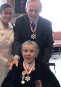 An elderly Bertha sits in a wheelchair wearing the Order of Canada Award medal with Prime Minister Jean Chretien standing behind her, a young lady is shown standing to his left.