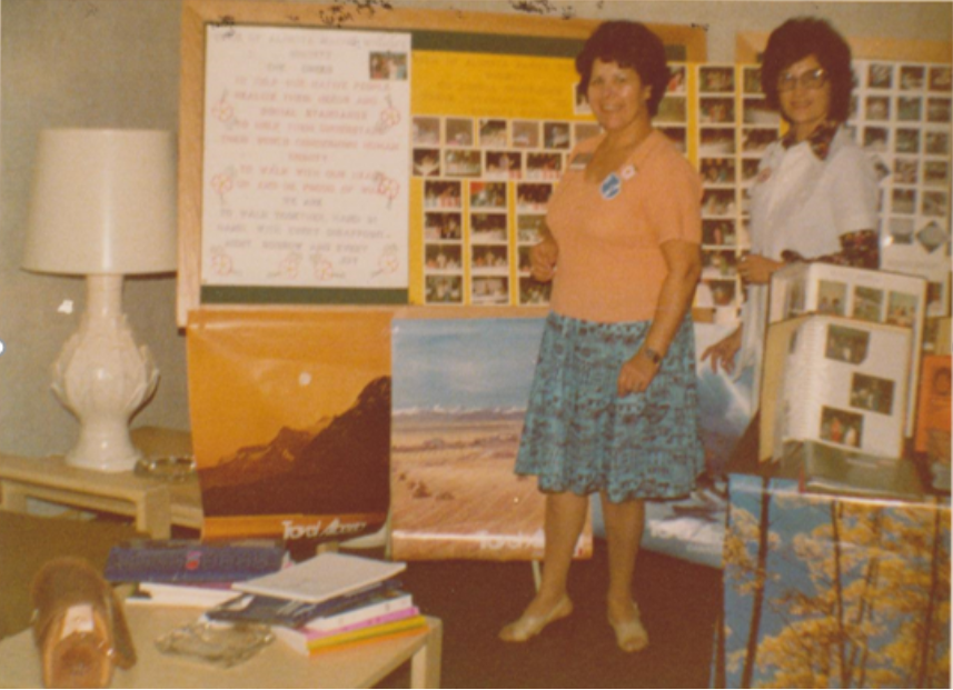 A middle-aged Bertha stands beside another woman in front of handmade photo collages and landscape paintings.