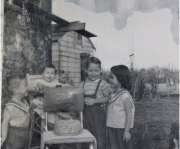 A black and white photo of four young children gathered smiling and standing around a birthday cake, the youngest sitting in a wooden highchair, a house and shed sitting in the background.