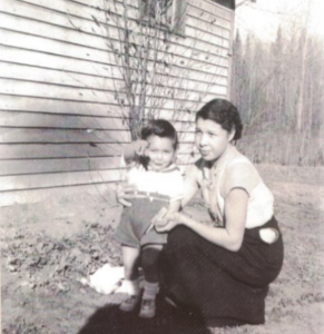 A young Elsie Cardinal kneeling next to her oldest son, the toddler is pointing toward himself, the background has a wooden structure in a treed yard.