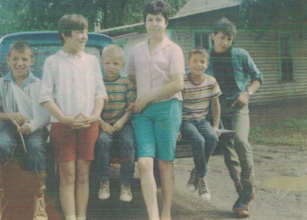 Elsie Cardinal as a young mother standing with one daughter and son, three boys sitting on the tailgate of a blue truck in the front yard of a house.