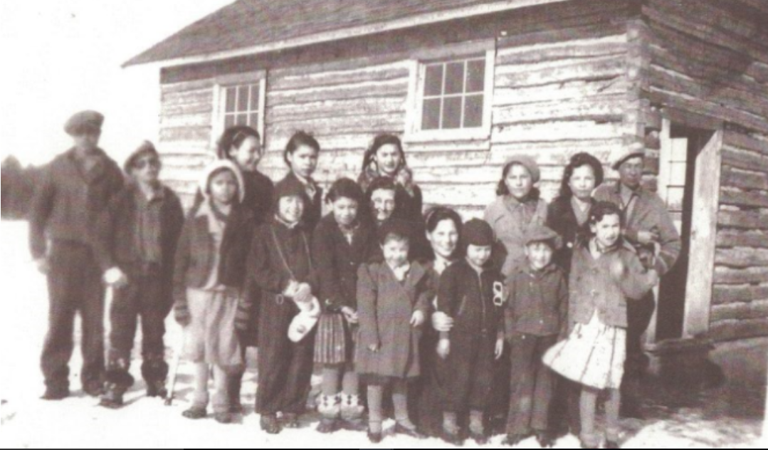 Black and white picture group of adults and children wearing winter clothing in front of a wooden building with snow on the ground.