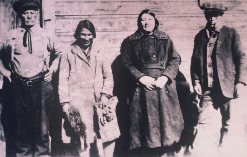 A historic photo of four people, two men and two women, standing in front of a wooden house, the second person is a young Mary-Rose.