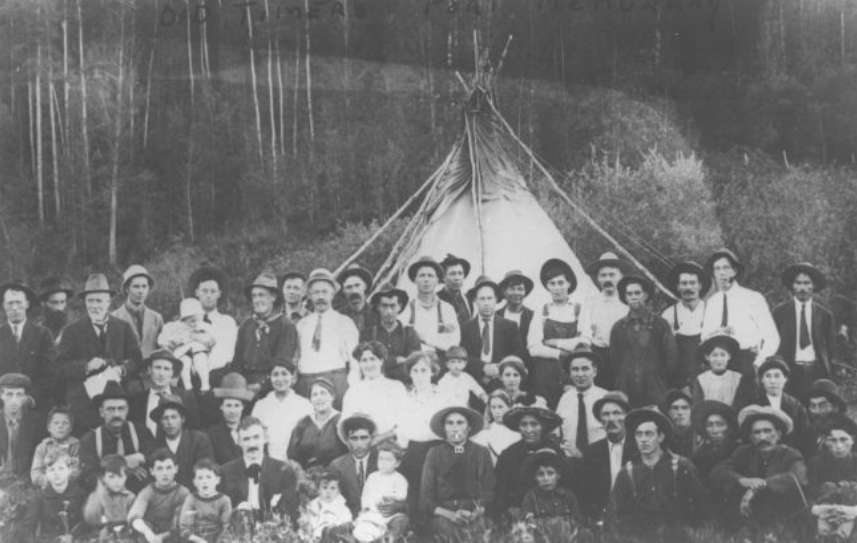 Black and white photo of a large group of people standing in front of a teepee looking straight ahead some are sitting down with young children while some are standing.