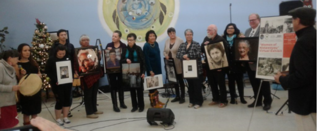 Women and men stand together in front of a photographer holding framed portraits of ten Indigenous women.