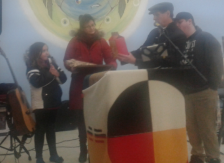 A woman and child are presented with a gift by a man with a man behind him, a podium sits in front of them covered by a quilted blanket, a colourful mural is shown on the wall in the background.