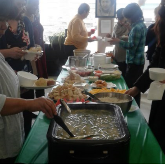 A group of people around a table with different types of food, soup, bannock, fruits along both side of the table with a green table plastic cloth.