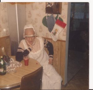 A male mummer in a nightgown and wearing a lampshade on his head sits at a table having a drink