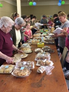 Baked goods are displayed on a table surrounded by customers waiting for the start of a bake sale