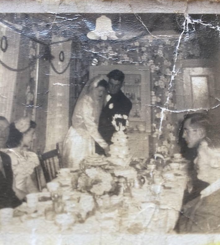 the bride and groom are shown cutting the wedding cake at Alice Drake's wedding supper in Oderin. guests and members of the bridal party look on.