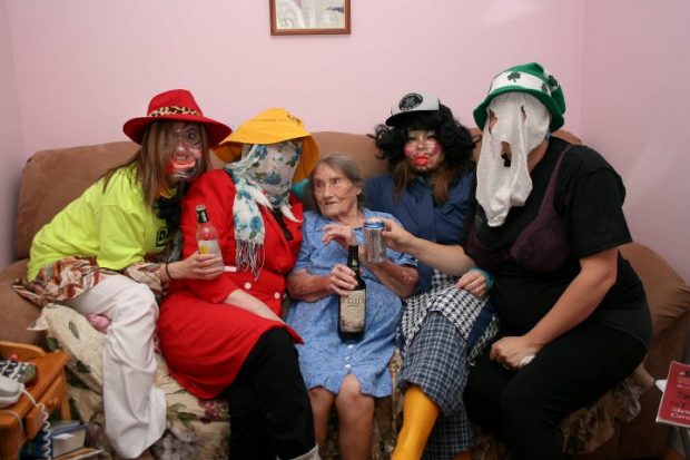 Four Mummers having a nip of rum sit with an elderly woman on a crowded couch