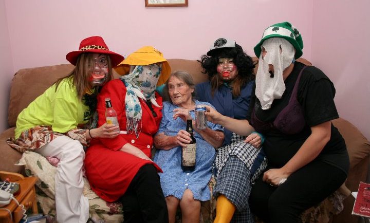 Four Mummers having a nip of rum sit with an elderly woman on a crowded couch