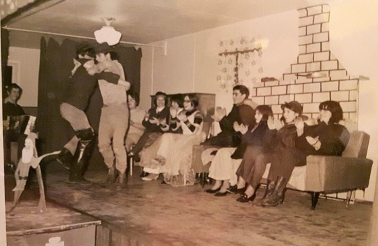 Two young men dance while other young people sit in the background and clap. another young man plays the accordion.
