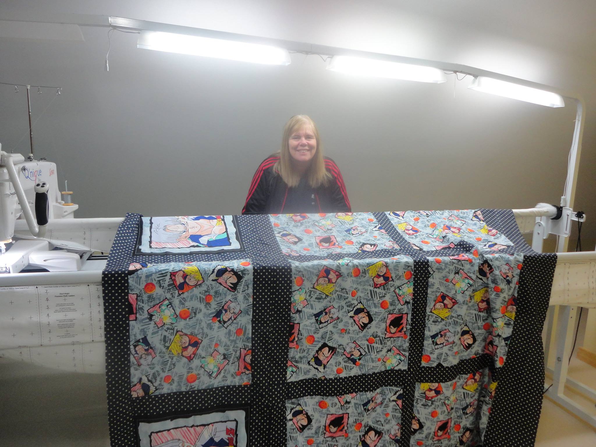 a woman stands behind a quilting machine displaying a quilt she is working on