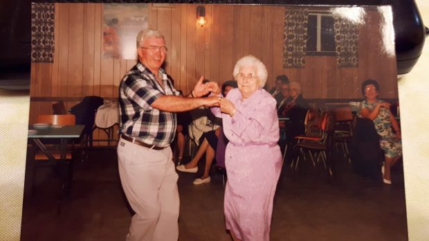 A man and an elderly woman in a pink dress dance in a community hall