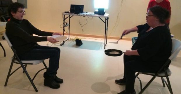 One woman tosses a pancake from a frying pan while another woman tries to catch it on a plate