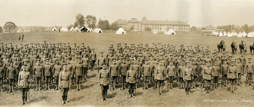 Photo d’un grand groupe de soldats, tous des hommes, en premier plan. Des tentes, des chevaux et un bâtiment à l’arrière-plan.