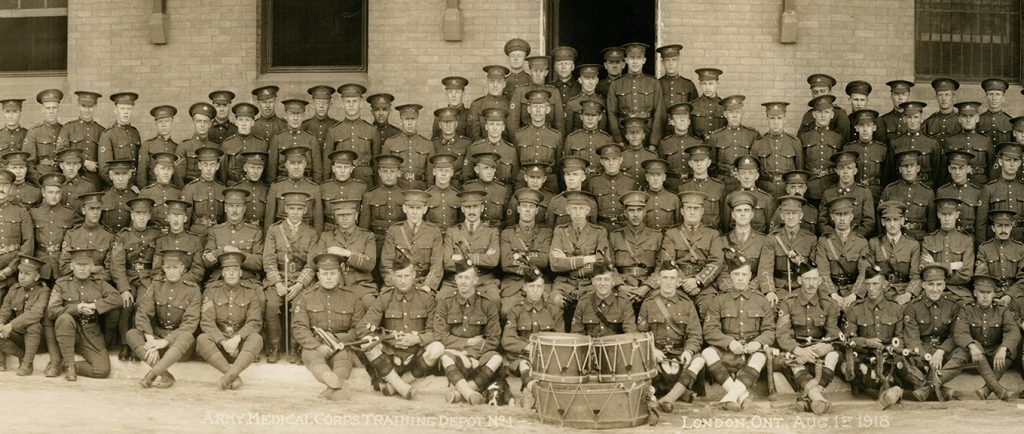 Photo d’un grand groupe de soldats, tous des hommes, assis et debout. Trois tambours en avant-plan et des détails de bâtiments à l’arrière.