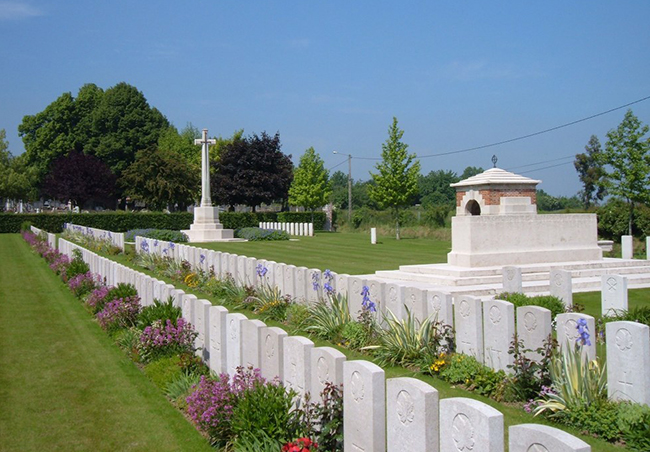 Photograph of a military cemetery with headstones in 2 rows surrounded by flowers. A large cross and the back of a munument are shown in the background.