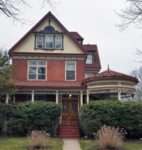 Une maison en briques rouges à deux étages et demi pourvue d’un porche et d’une véranda circulaire. Devant la maison, on peut voir un sentier qui mène à la porte d’entrée et de la verdure.