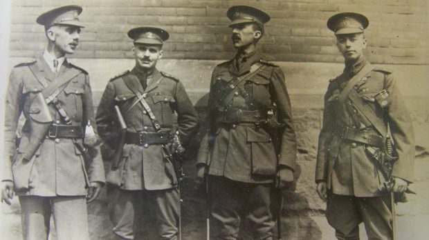 Four males, military officers standing and talking in front of a building. Second from right holds his hands behind his back. They all have pistol holsters and swords.