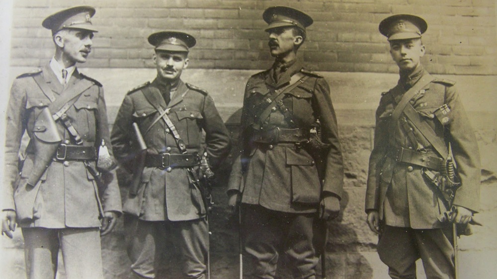 Four males, military officers standing and talking in front of a building. Second from right holds his hands behind his back. They all have pistol holsters and swords.