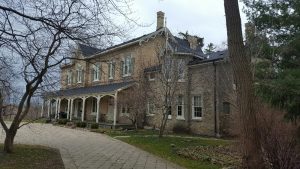 Une grande maison de 2 étages dans un jardin. La maison est en brique jaune, aux jolis motifs décoratifs en bois blanc; l'entré principale se trouve sious un large porch qui couvre toute la façade. Un bâtiment adjacent, plus petit se voit derrière un petit arbre.