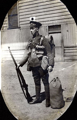 The image of a soldier standing in an oval mat. He is holding his rifle with his right hand. A duffel bag is beside his left foot. A building is seen behind.