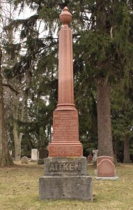 Un monument funéraire en forme d'obélisque, une structure en marbre rouge surmontant une base en granite gris avec l'inscription