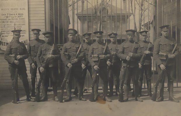 Ten soldiers, all wearing a peak hat and holding rifles. They stand in front of a grill gate, a fragment of a sign for a theatre can be seen on the right.
