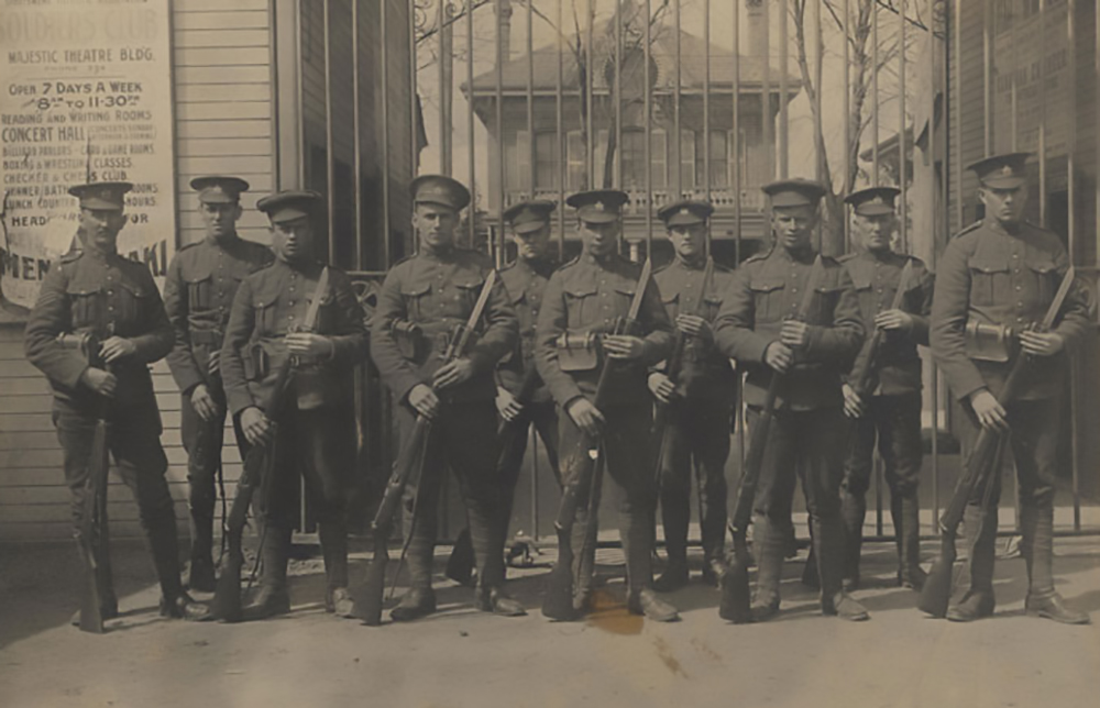 Ten soldiers, all wearing a peak hat and holding rifles. They stand in front of a grill gate, a fragment of a sign for a theatre can be seen on the right.