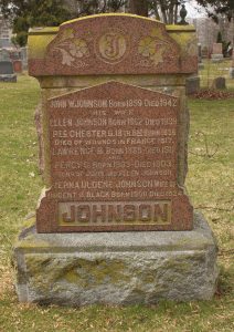 A head stone in the shape of a sarcophagus. The upper part is decorate with a plinth that has carved flowery ornaments and the initial "J" in Gothic fonts..