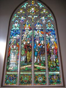 A monumental stained glass in a church, formed of 4 main panels, surmounted with Gothic ornaments closed in a pointed arch (ogive).