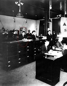 Photograph of an office with tall desks, 9 males and 1 female workers.