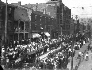 A military parade on the street of a city. The parade is lead by a carriage with 2 horses, the soldiers are marching in 2 rows. Crowds watch on the sidewalk.