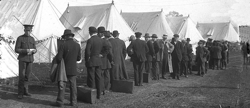 Photographie d'une file d'attente formée des hommes habillés en tenue de civil, certains ont des valises sur terre. On peut voir un soldat tenant un morceu de papier et des tentes blancs.