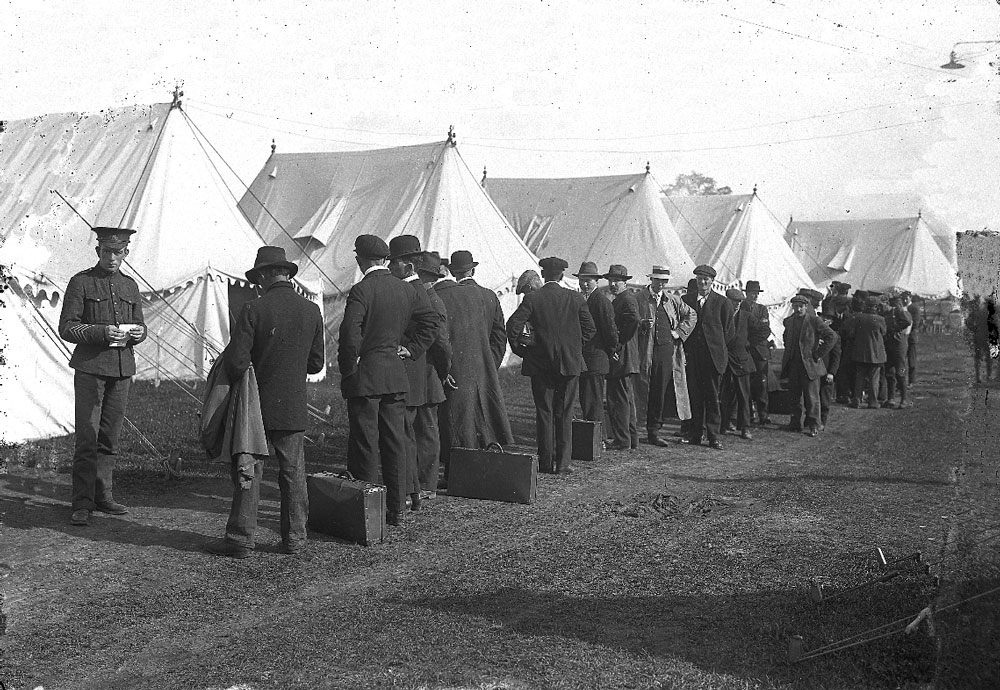 Photograph of a line up of men in civilian dress, some have suitcases on the ground. A soldier holding a piece of paper and white tents can be seen.