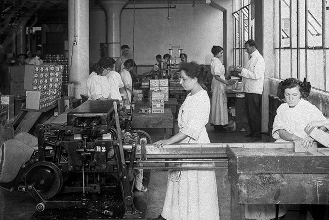 Photographie en noir et blanc dans une usine; en premier plan 2 femmes travaillent derrière une machine industrielle. Plusieurs autres personnages en arrière-plan, femmes et hommes.