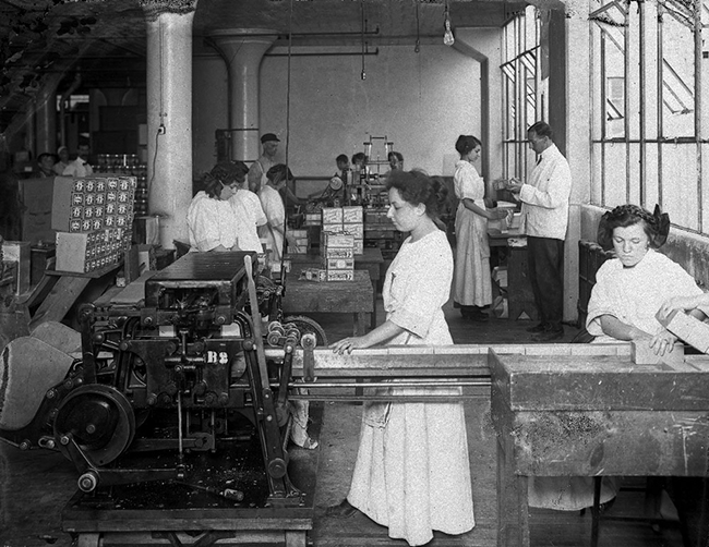 Black and white photograph in a factory; in front, 2 women are working behind an industrial machine. Other characters, women and men, in the background.