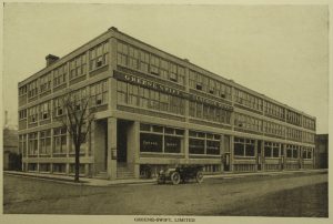 Le bâtiment d'une usine, avec une voiture et une bicyclette devant. On voit une affiche avec l'inscription