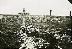 A photograph showing a graveyard with many crosses. In the foreground 3 crosses, the first is white in crowns a grave that has flowers on it.