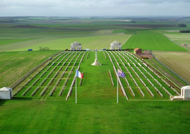 Photograph of a military cemetery ith many rows of headstones in a landscape. French and Australian flags are in front.