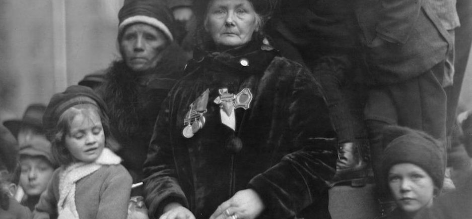 Photo représentant une femme endeuillée, arborant des médailles et tenant un sac-à-main; elle est entourée d’enfants et d’une autre femme endeuillée.