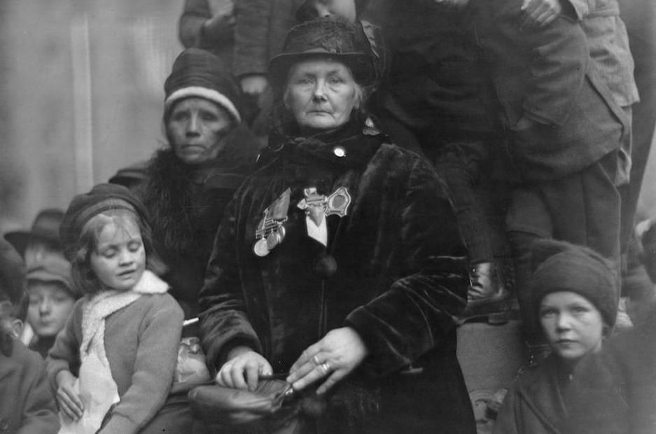 Photograph featuring a women in mourning who has medals on her chest and holds a purse; she is surrounded by children and another women in mourning.