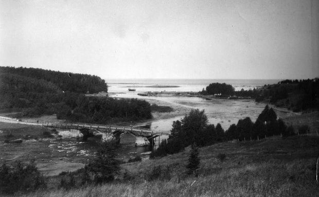 Photograph of the old wooden Bridge at Grand-Métis circa 1925. The bridge is located about 1 km from the mouth of the Metis River. Both sides of the river present a forest landscape and wharf. A schooner is moored at the mouth of the river and offers a glimpse of the expanse of the St. Lawrence River.